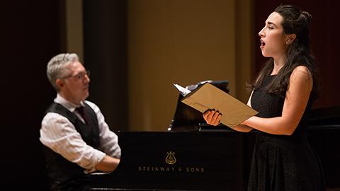 An opera singer on stage in a black dress, holding a score in her hands. Behind her is a pianist, sitting at a grand piano