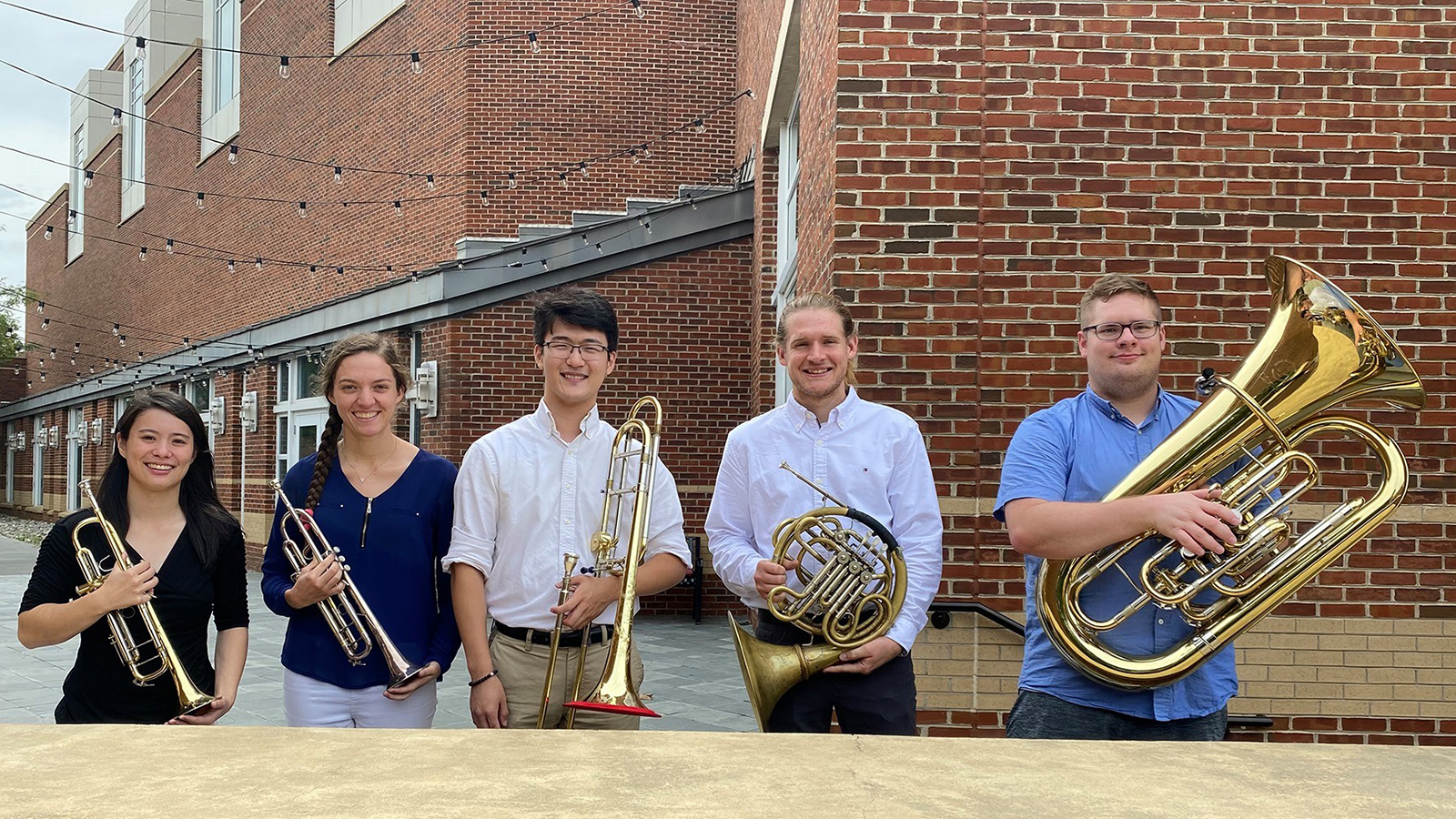  The five members of the inaugural UMD Terrapin Brass ensemble standing outside The Clarice in a courtyard holding their brass instruments while smiling.