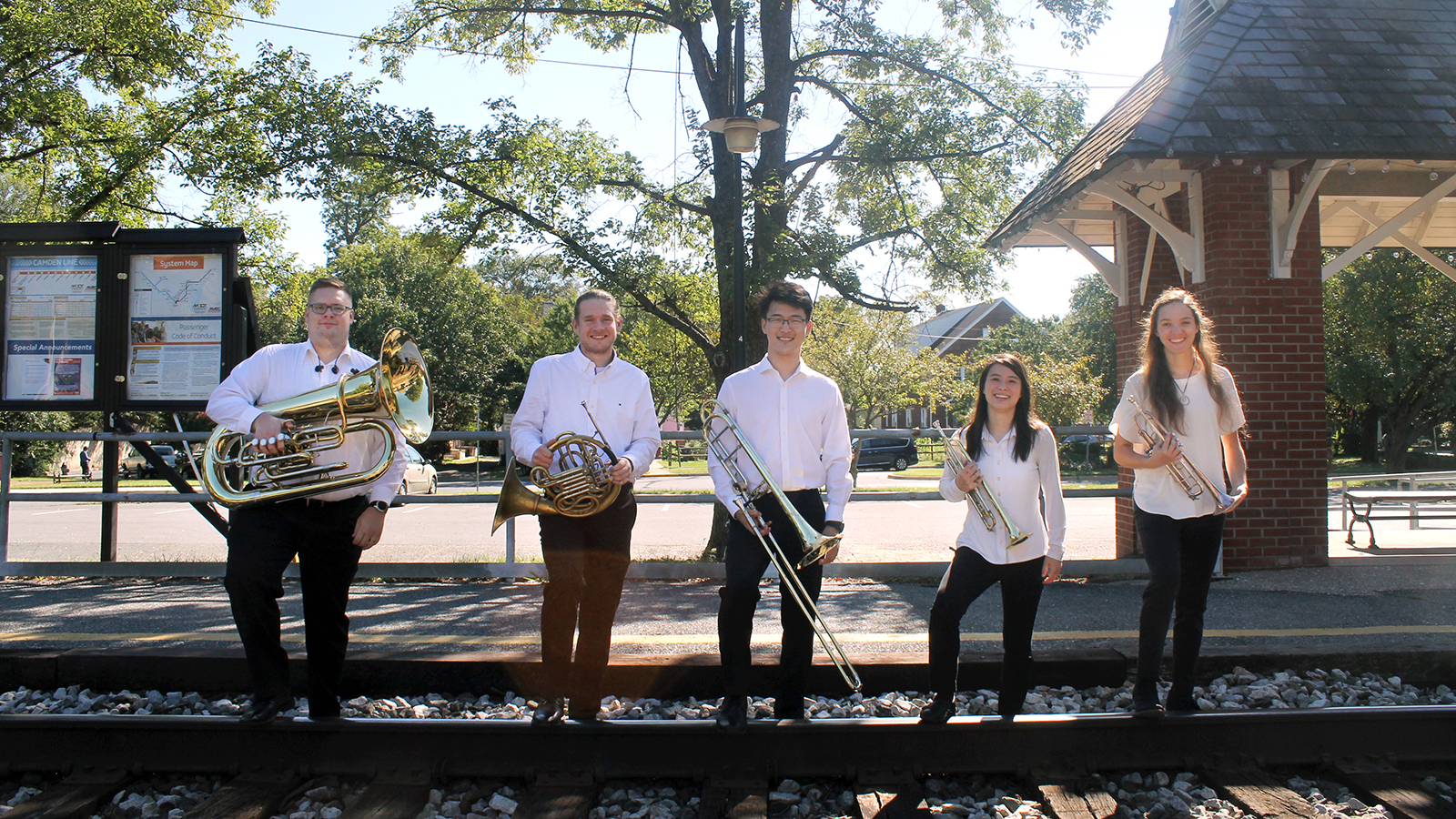  The five graduate students of Terrapin Brass stand on the train tracks holding their brass instruments and smiling.
