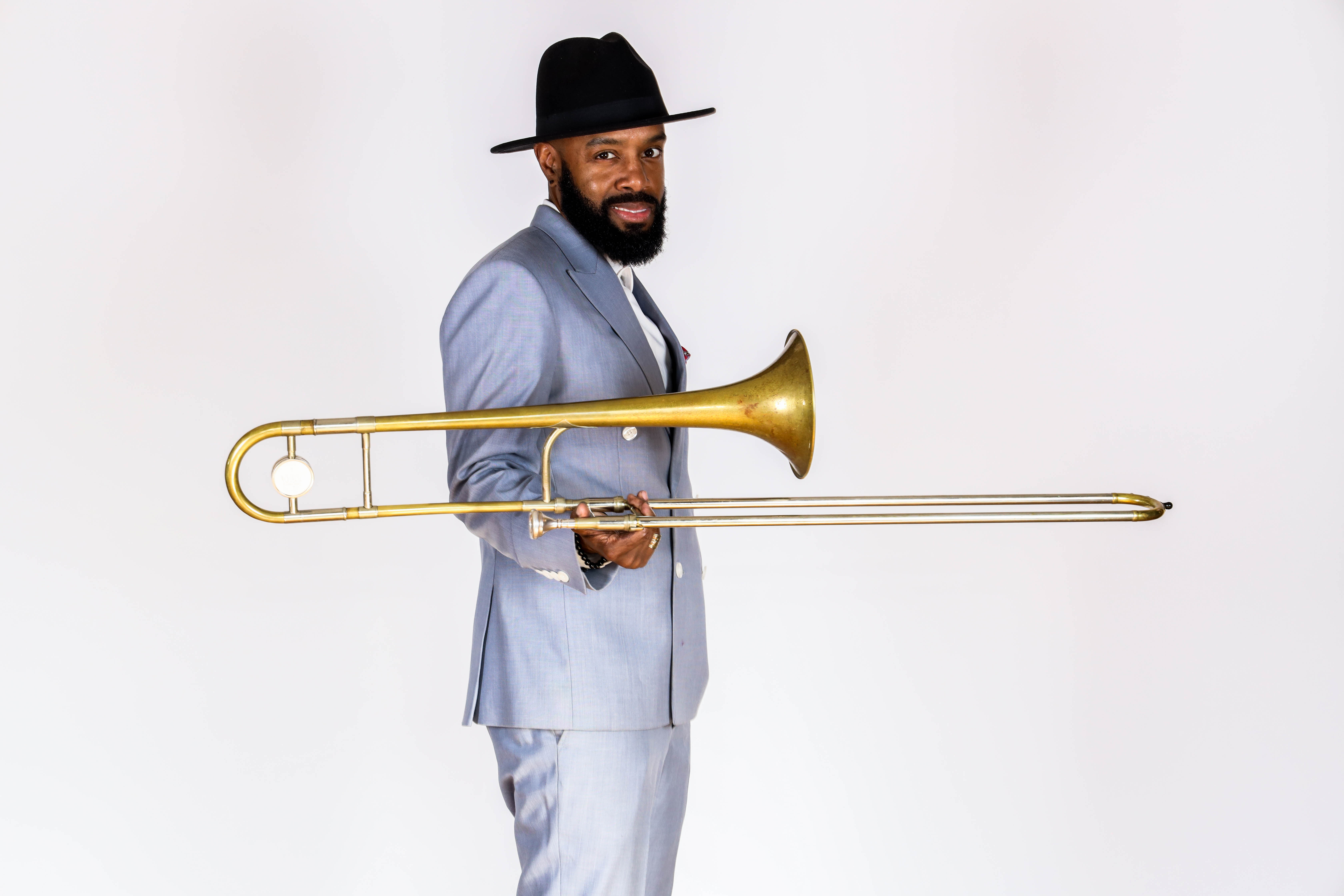 Mark Williams, dressed in a light blue suite against a white backdrop, holding his trombone out in front of him.