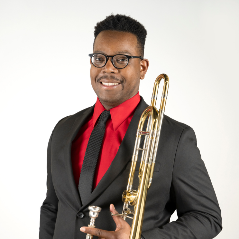 Assistant Trombone Professor Myles Blakemore. He is dressed in a black suit with a red button up shirt, and his holding his trombone.