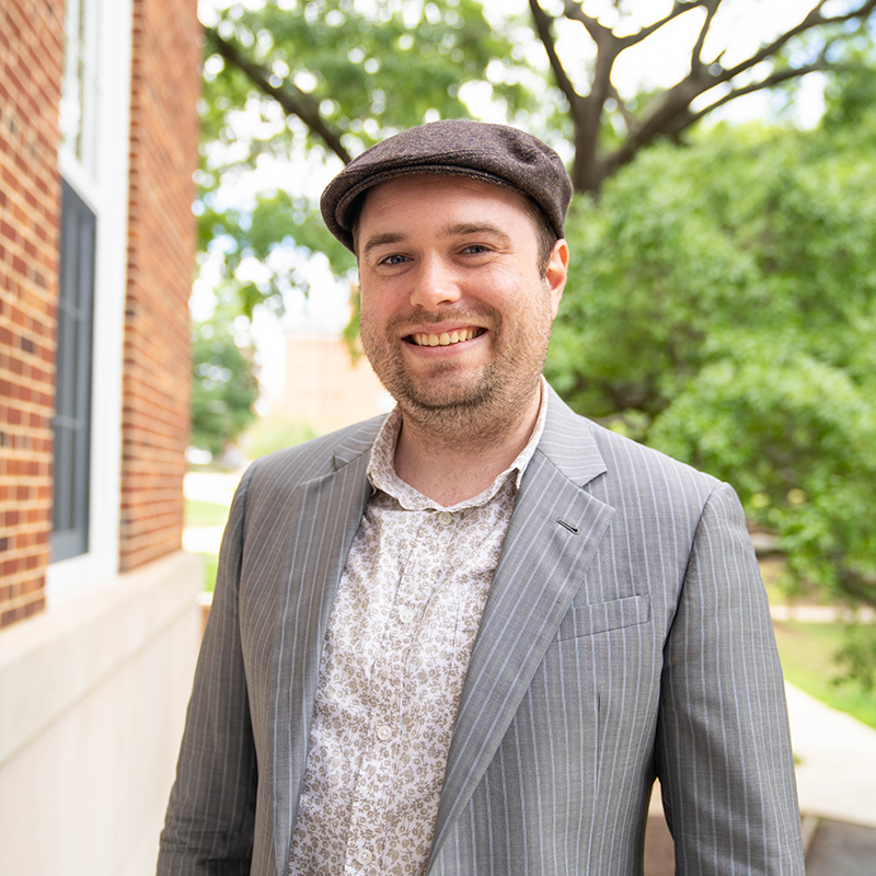 Assistant Clinical Professor Johannes Visser. He is wearing a brown newsboy cap, floral patterned shirt, and a light gray jacket with stripes. 