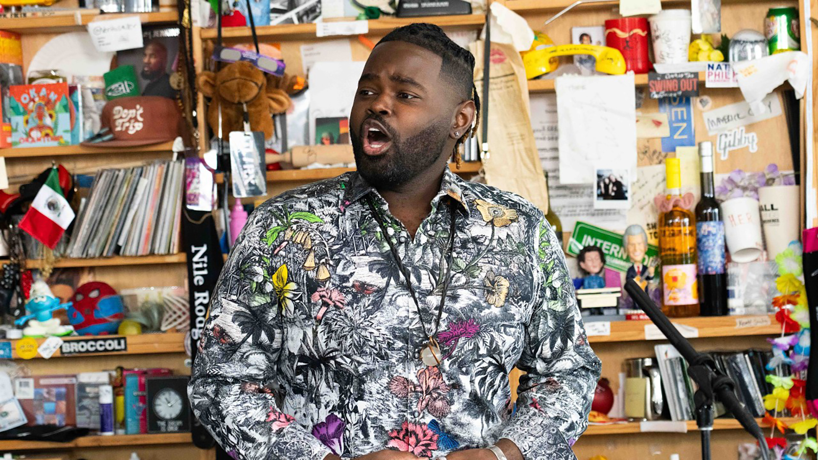 Associate Professor of Voice John Holiday. He is on the set of NPR's Tiny Desk, with a very cluttered background. He is wearing a photorealistic tropical print suit, and he is singing.