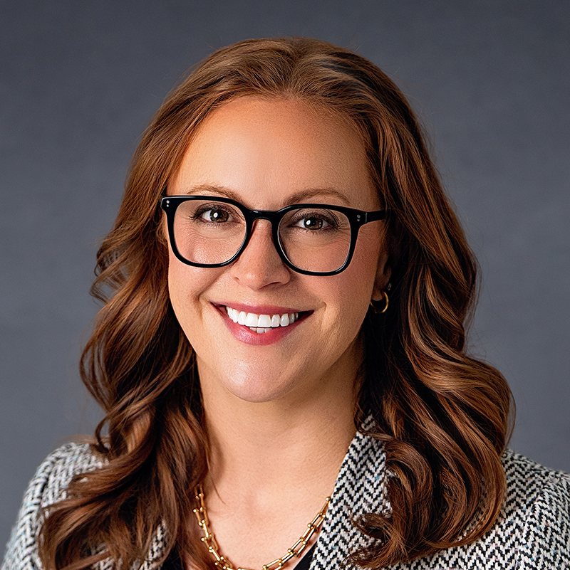 Stephanie Prichard. She has brown hair with reddish highlights, glasses, and is wearing a black and white patterned jacket. She is smiling at the camera.