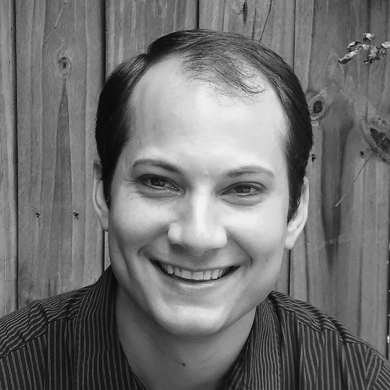 J Bradley Baker. The image is in black and white and Baker is posed against a wooden backdrop