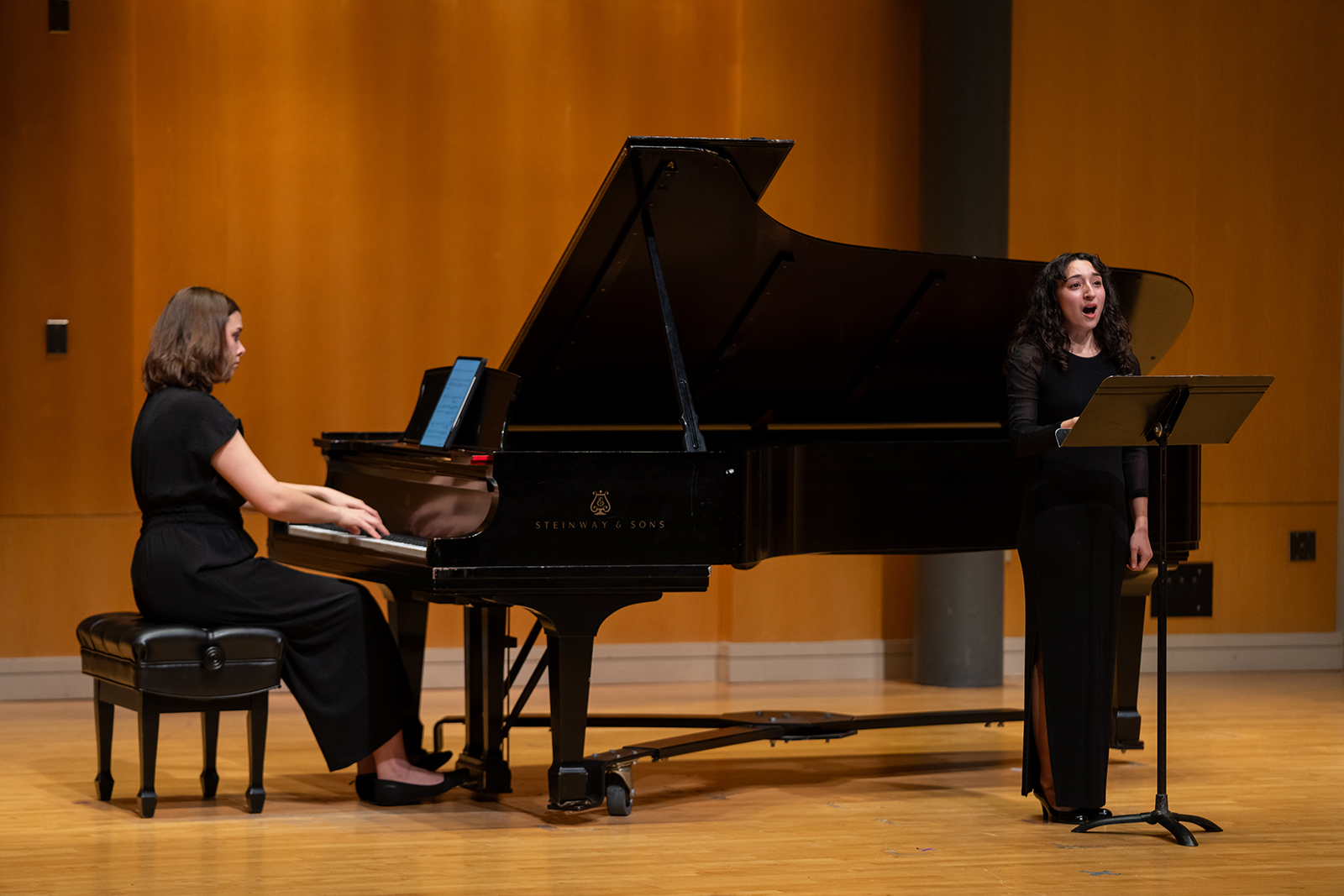 A vocalist performs on stage. In front of her is a music stand, and behind her is a pianist accompanying her.