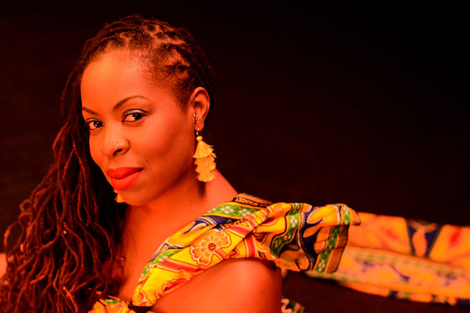 Johnaye Kendrick Poses for a headshot. She is posed against a black background and has her braids covering one shoulder. She is wearing yellow earrings and a yellow patterned dress.