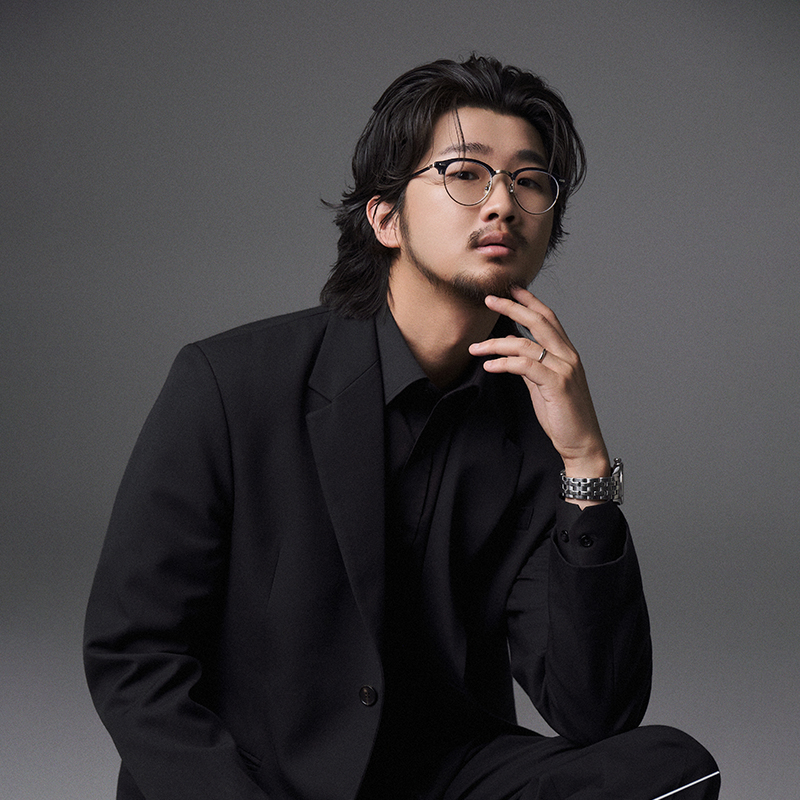 A headshot of Eric Kim. He is wearing concert black and is posing with his elbow on his knee and his fingers positioned on his chin.