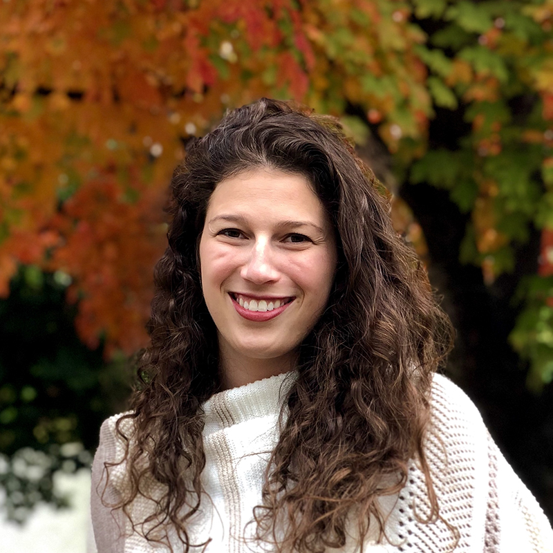 Nicole Steinberg poses for a headshot. She is a white, brunette woman wearing a knitted shawl and smiling at the camera. In the background is fall foliage.