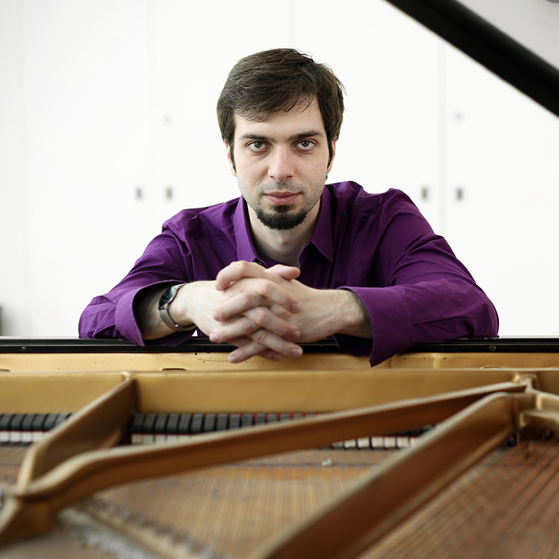 Chris Ziemba sits at a piano, his hands draped over the the top of the tuning pins. He is wearing an eggplant colored shirt, and looking directly into the camera.