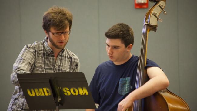 Photo of high schooler, holding an upright bass, receiving instruction on a piece of music