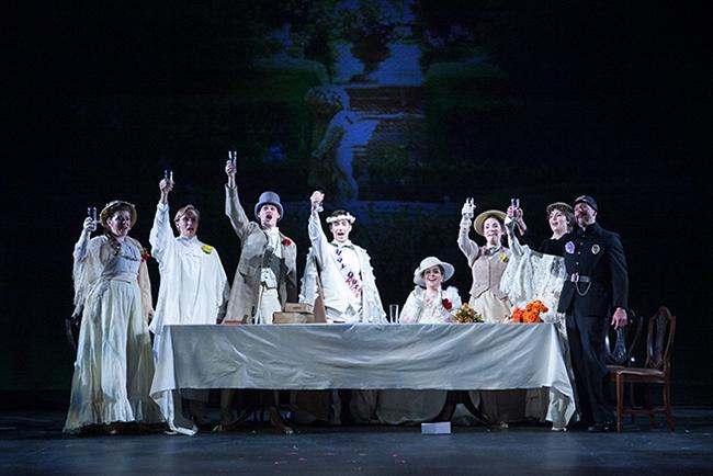 A group of costumed figure on stage, raising glasses as if toasting. Each person wear an entirely white costume, save for a singular person dressed as a police officer, who is dressed in all black.